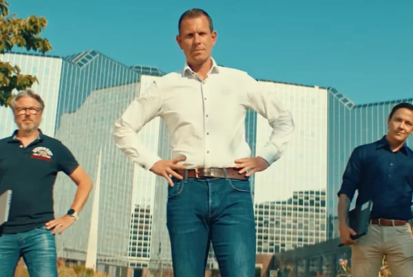 Drie mannen, waarvan de middelste een wit overhemd draagt, staan voor groot gebouw met glazen pui.